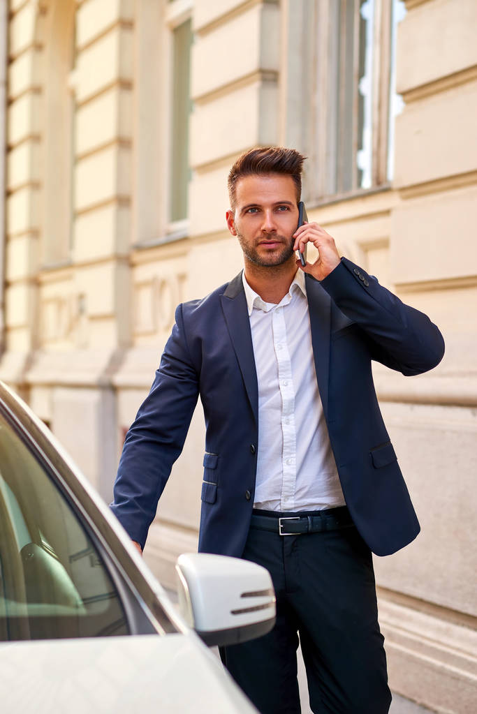 Nuori liikemies kävelee autolleen ja puhuu puhelimessa.
 - Valokuva, kuva