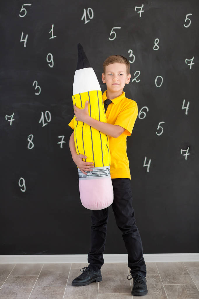 Lindo chico guapo de la escuela en camiseta amarilla corbata y botas elegantes casuales de pie cierre a la pizarra negra con números y la celebración de enorme lápiz de juguete suave y sonriente
 - Foto, imagen
