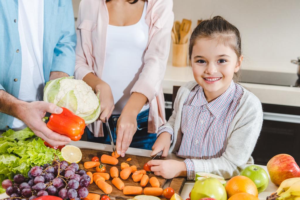 imagem cortada da família na cozinha, criança pequena com a mãe cortando legumes e olhando para a câmera
 - Foto, Imagem