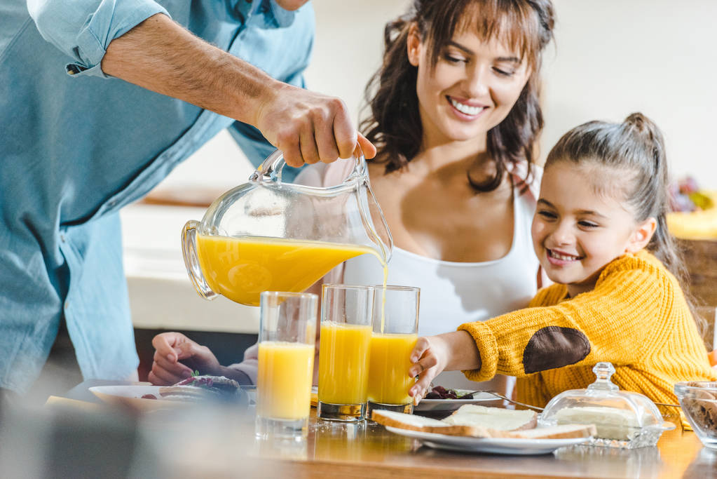 ευτυχισμένη οικογένεια στο τραπέζι, ο άνθρωπος ρίχνει χυμό σε ποτήρια με γυναίκα και παιδί στην κουζίνα - Φωτογραφία, εικόνα