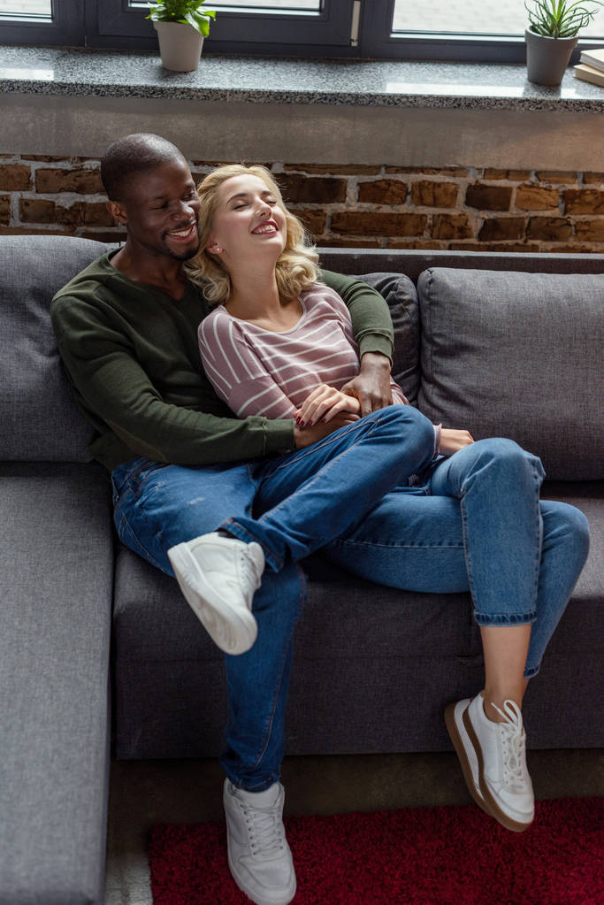 high angle view of happy african american man hugging girlfriend while resting on sofa together - Photo, Image