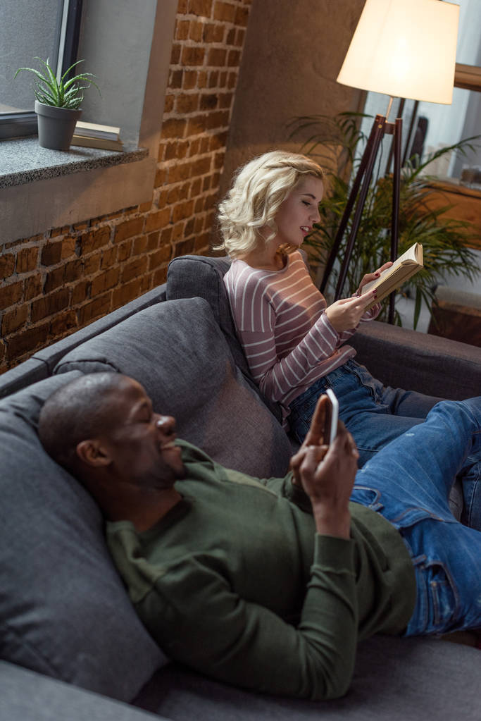 Afro-Amerikaanse man met smartphone terwijl Kaukasische vriendin lezen boek op de Bank thuis - Foto, afbeelding