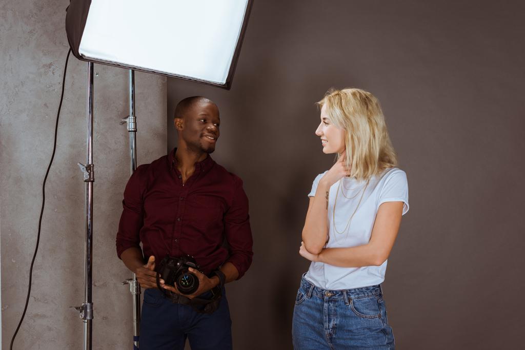 cheerful multiethnic photographer and model looking at each other while having photoshoot in studio - Photo, Image