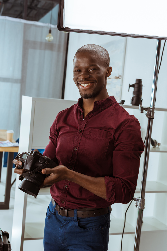 Porträt eines fröhlichen afrikanisch-amerikanischen Fotografen mit einer Fotokamera in der Hand, die auf die Kamera im Studio blickt - Foto, Bild