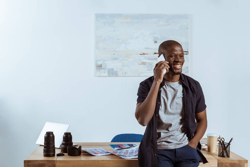 portret uśmiechający się amerykański fotograf mówi na smartfonie w miejscu pracy w studio - Zdjęcie, obraz