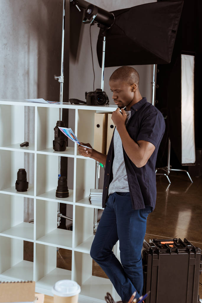 vista lateral del fotógrafo afroamericano enfocado mirando papeles en la mano en el estudio
 - Foto, imagen
