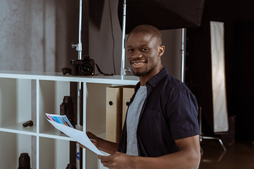 podobizna veselá afroamerické muže s papíry v ruce při pohledu na fotoaparát ve studiu - Fotografie, Obrázek