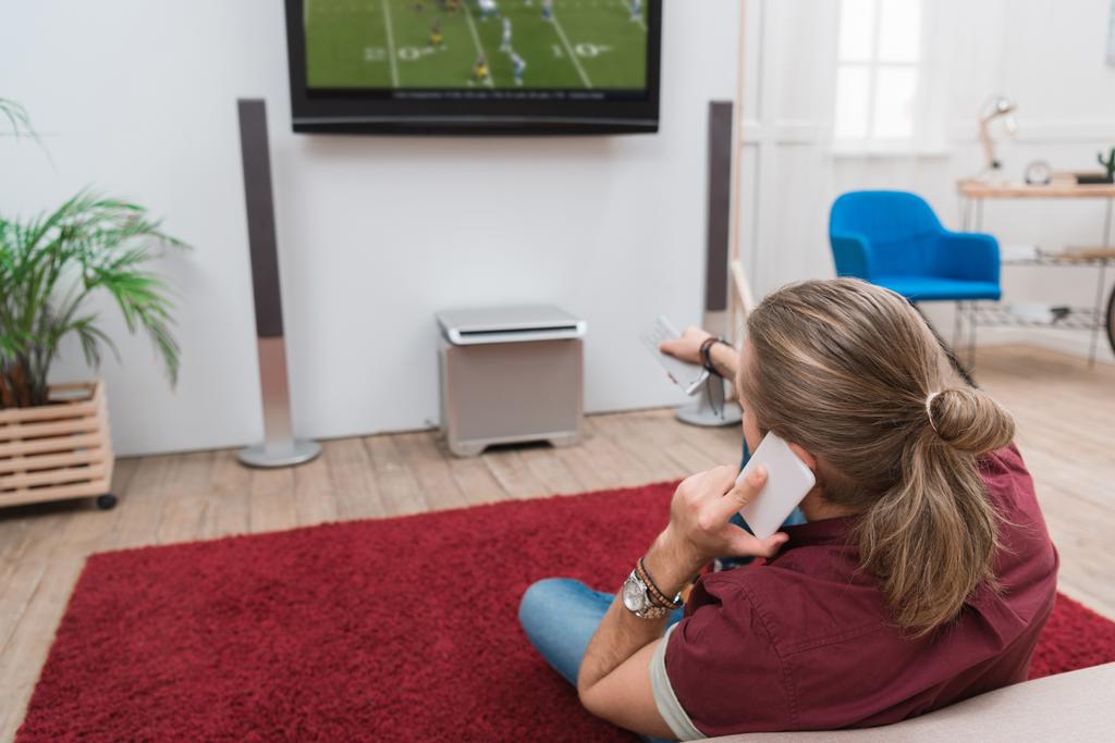 Вид сзади человека, разговаривающего по смартфону во время просмотра футбольного матча дома
  - Фото, изображение