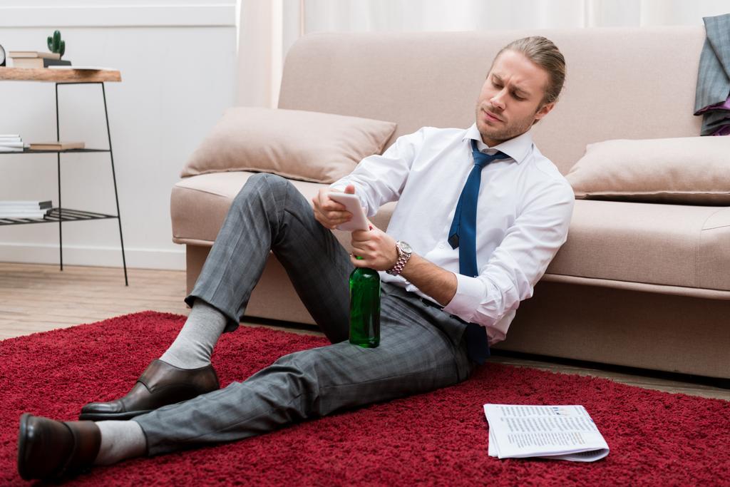 Knappe man, zittend op een vloer in de woonkamer met smartphone en flesje bier - Foto, afbeelding