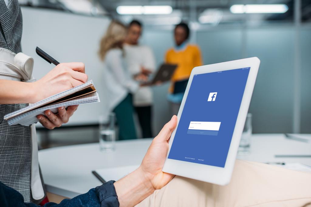 Geschäftsmann hält Tablet mit Facebook am Bildschirm im modernen Büro, während Kollege Notizen macht - Foto, Bild