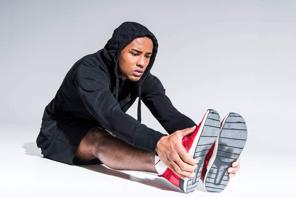 jeune sportif afro-américain en baskets étirant les jambes sur gris
 - Photo, image