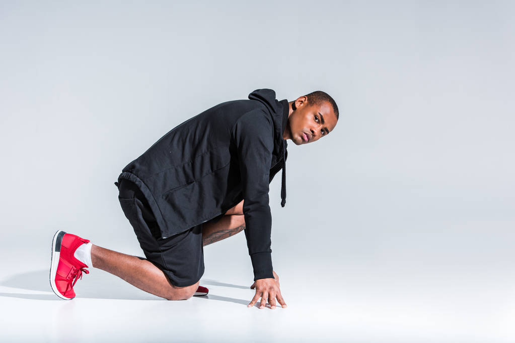 vue latérale du jeune sportif afro-américain en sweat à capuche prêt à courir et regardant la caméra sur gris
 - Photo, image