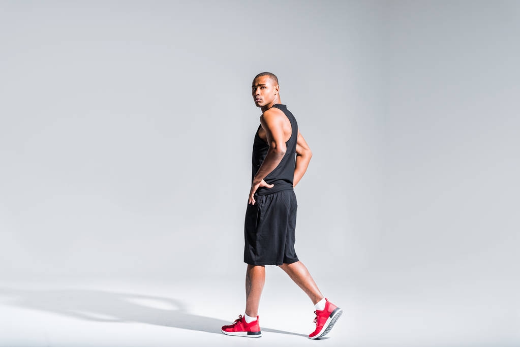 молодой африканский американский спортсмен с руками на талии ходит и смотрит в камеру на сером
 - Фото, изображение