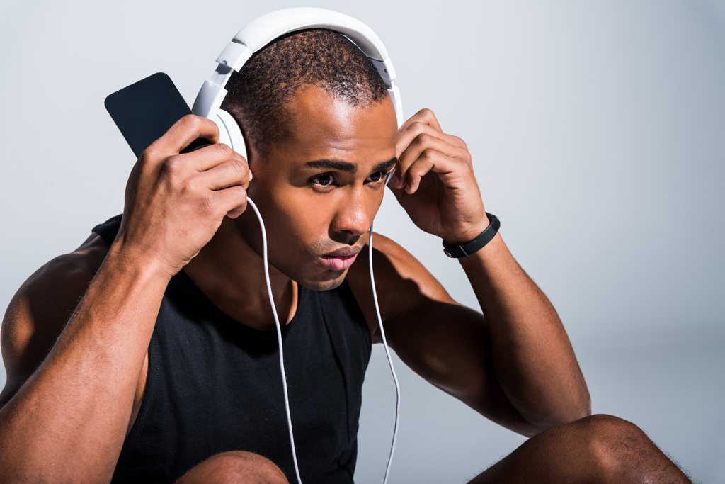 jeune sportif afro-américain dans les écouteurs à l'aide d'un smartphone et en regardant loin sur gris
 - Photo, image