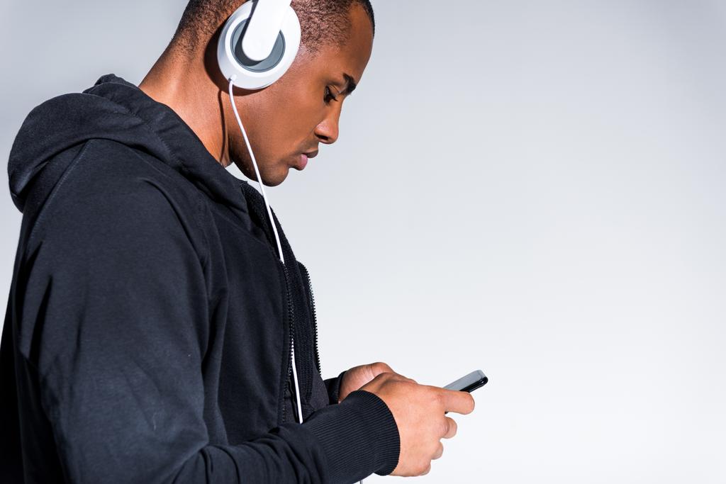 Μερική άποψη του νεαρός αφρικανική αμερικανική άνθρωπος στα ακουστικά χρησιμοποιώντας το smartphone που απομονώνονται σε γκρι  - Φωτογραφία, εικόνα
