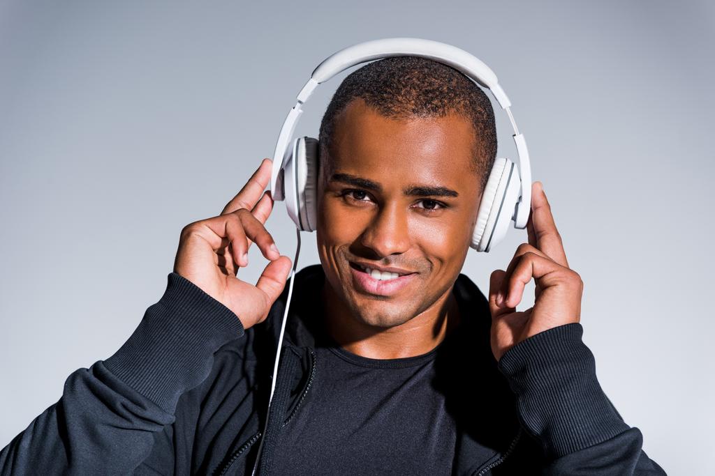 csinos fiatal afro-amerikai sportoló hallgat zenét a fejhallgatót, és mosolyogva kamera elszigetelt szürke - Fotó, kép