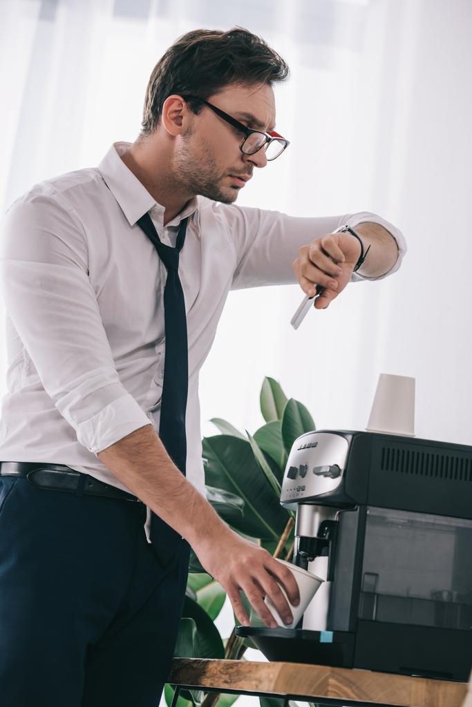 бизнесмен наливает кофе из офисной машины и смотрит на часы
 - Фото, изображение