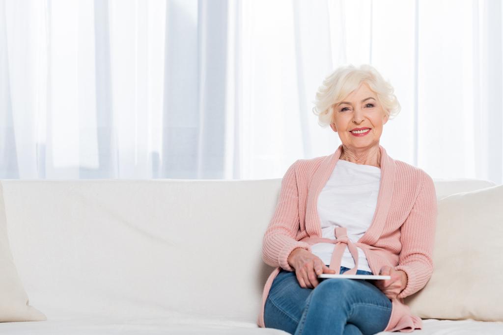 portrait of smiling senior woman with tablet in hands resting on sofa and looking at camera at home - Photo, Image