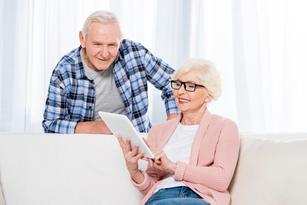Porträt einer Seniorin mit Tablet, während ihr Mann in der Nähe zu Hause steht - Foto, Bild