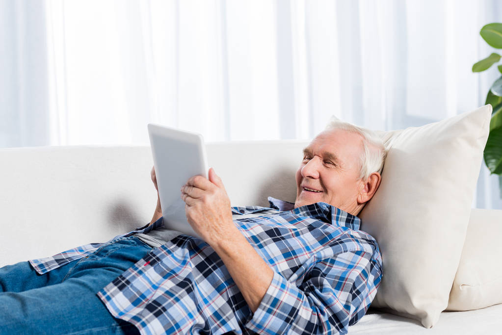 Seitenansicht eines fröhlichen Seniors, der auf dem Sofa liegt und zu Hause ein Tablet benutzt - Foto, Bild