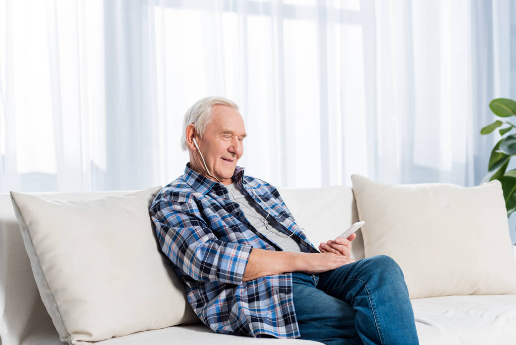 puolella näkymä hymyilevä vanhempi mies kuulokkeet älypuhelin lepää sohvalla kotona
 - Valokuva, kuva