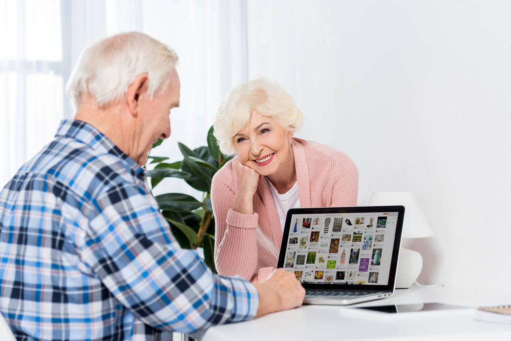 Porträt einer glücklichen Seniorin, die ihren Mann mit Laptop und Pinterest-Logo zu Hause betrachtet - Foto, Bild