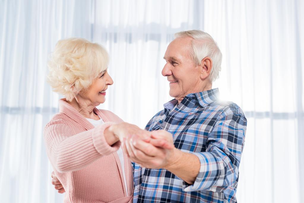 вид сбоку улыбающейся пожилой пары, танцующей вместе дома
 - Фото, изображение