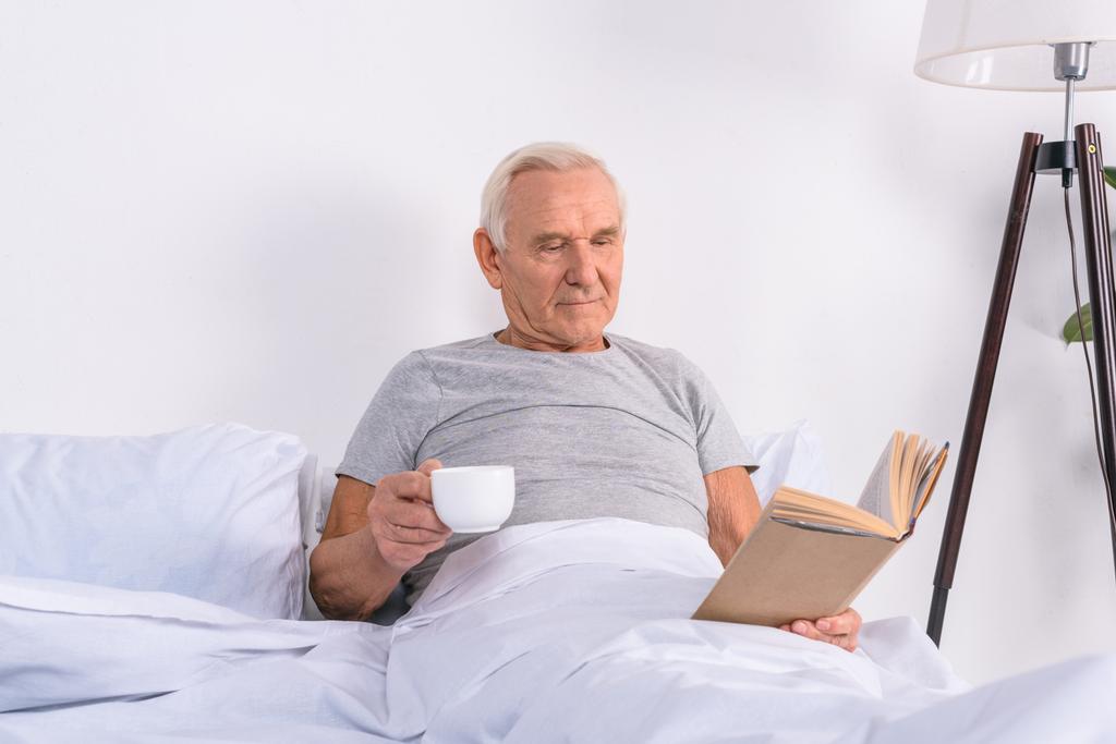 retrato de homem sênior com xícara de livro de leitura de café na cama em casa
 - Foto, Imagem