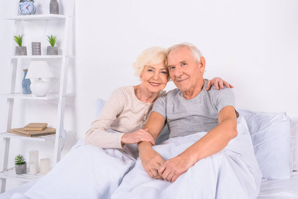 Portret van lachende senior paar rusten op bed en camera thuis kijken - Foto, afbeelding