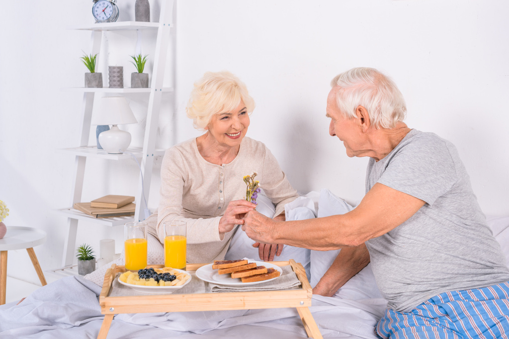 年配の男性が一緒に自宅のベッドで朝食をとりながら妻に花を提示 - 写真・画像