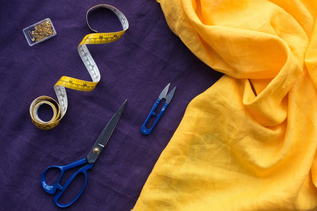 вид сверху на ножницы, измерительную ленту и коробку с булавками поверх фиолетовой ткани
 - Фото, изображение