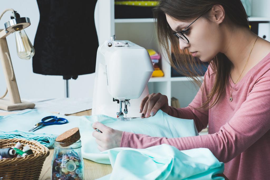 Zijaanzicht van jonge naaister met naaimachine op werkplek - Foto, afbeelding