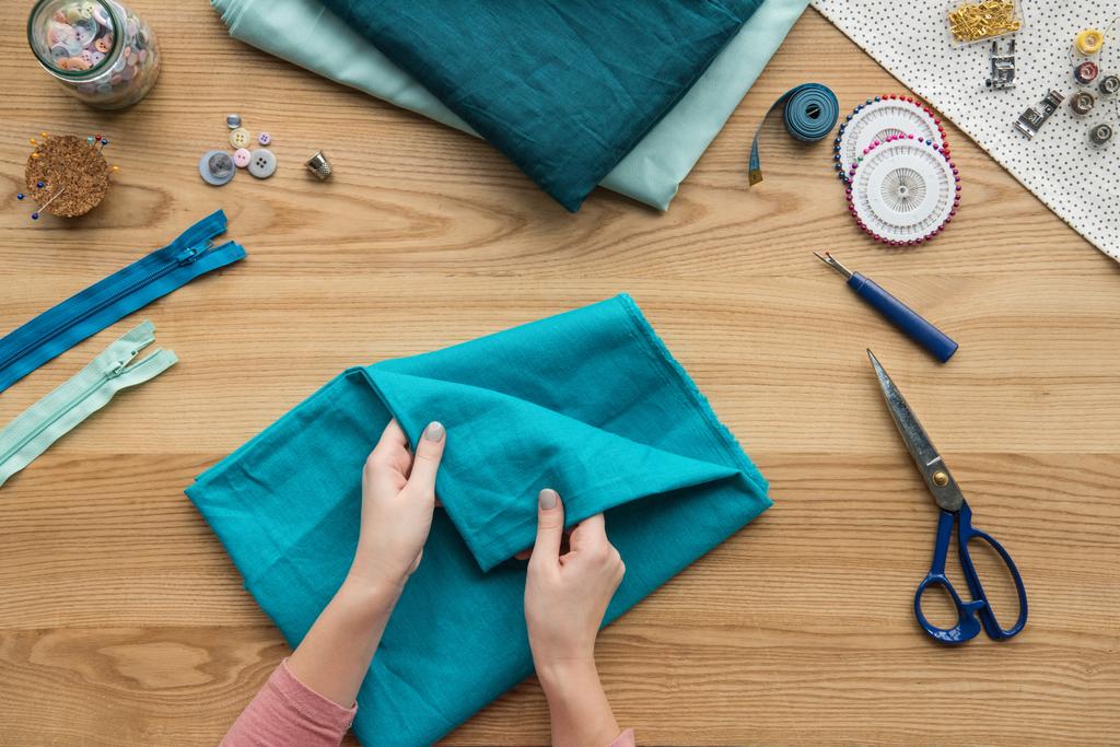 vista superior do tecido dobrável das mãos femininas cortadas no local de trabalho da costureira com tesouras e botões
 - Foto, Imagem