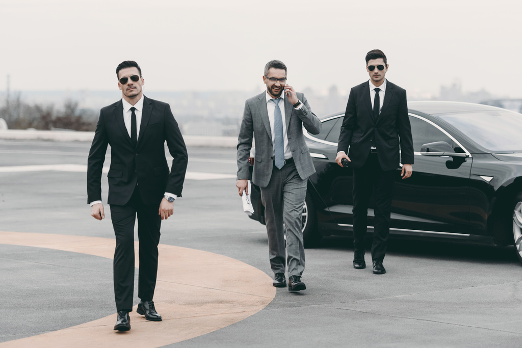 smiling businessman walking with bodyguards from car and talking by smartphone   - Photo, Image