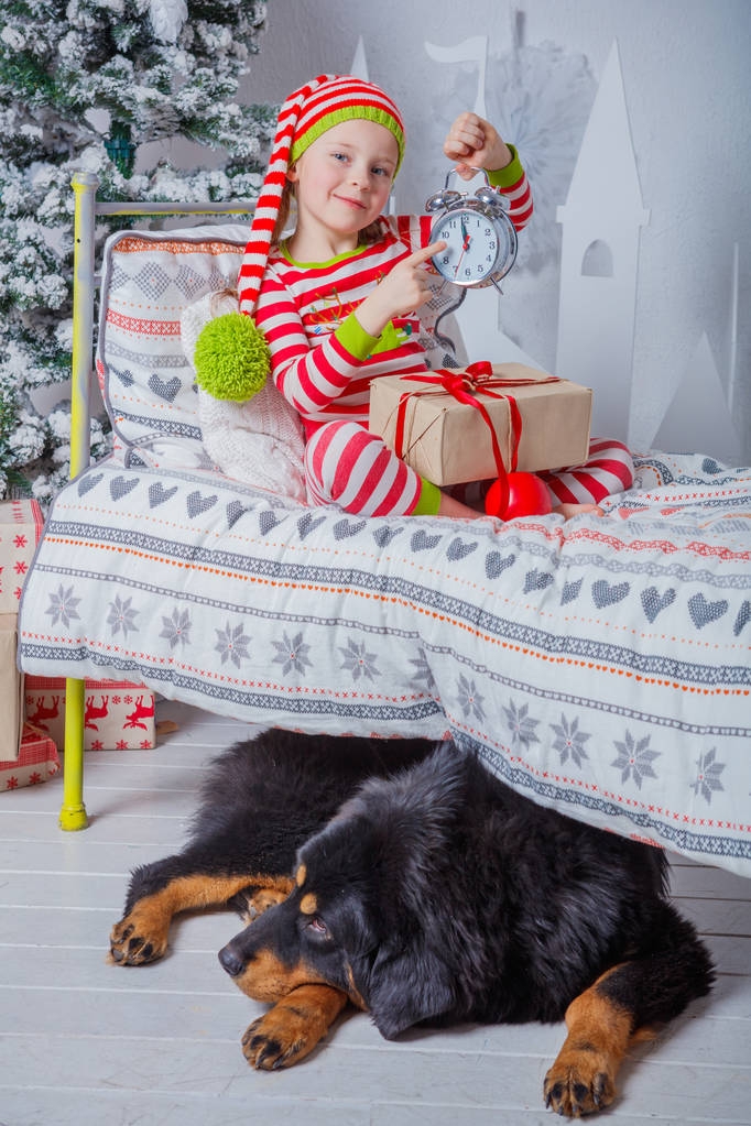 glückliches nettes kleines Mädchen in gestreiften Pyjamas sitzt im dekorierten Neujahrszimmer zu Hause. - Foto, Bild