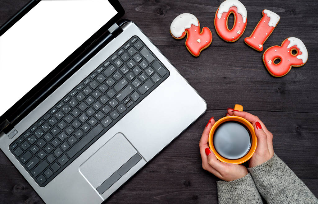 Ansicht von Frauenhänden mit Tasse heißen Kaffees, offenem Laptop mit weißem Bildschirm und Neujahrszeichen 2018 aus Lebkuchen, Kopierraum. flache Lage - Foto, Bild