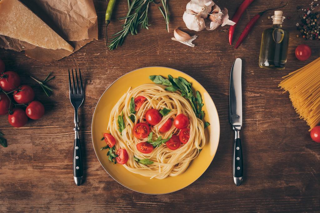 acostado plano con pasta italiana tradicional con tomates y rúcula en plato sobre mesa de madera con ingredientes
 - Foto, Imagen