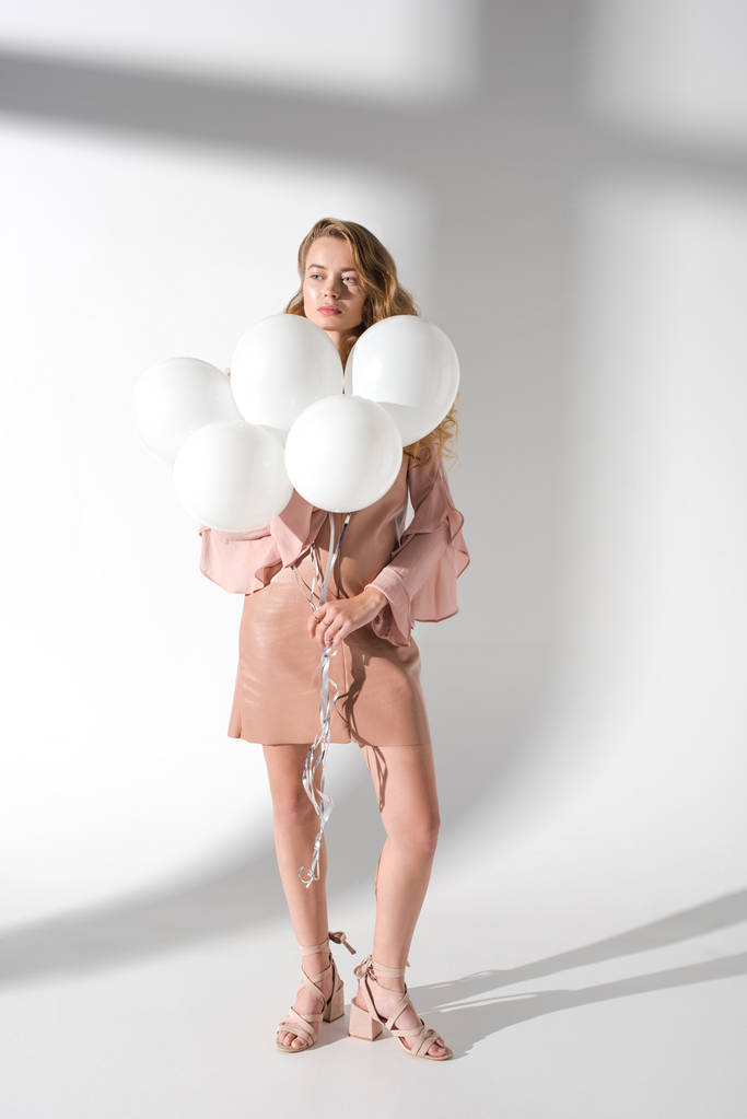 attraktive Frau in beigem Kleid mit Bündel weißer Luftballons und wegschauendem Blick - Foto, Bild