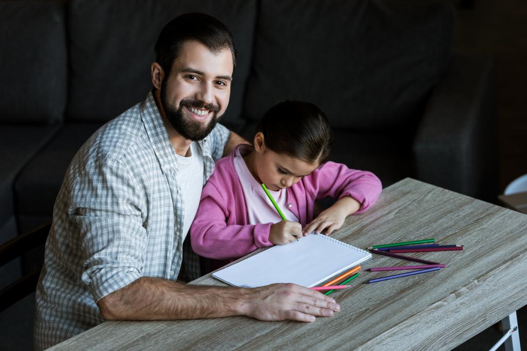 glücklicher Vater mit kleiner Tochter am Tisch sitzend und daheim in Sammelalbum zeichnend - Foto, Bild