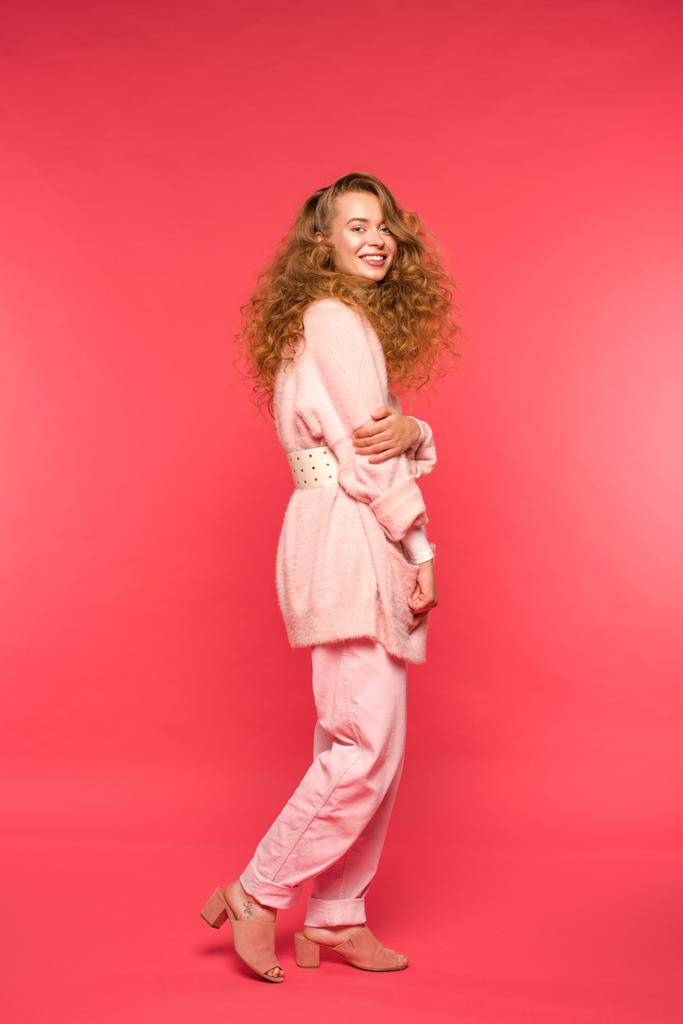 stijlvolle lachende meisje permanent in roze kleding en kijken naar camera geïsoleerd op rood - Foto, afbeelding