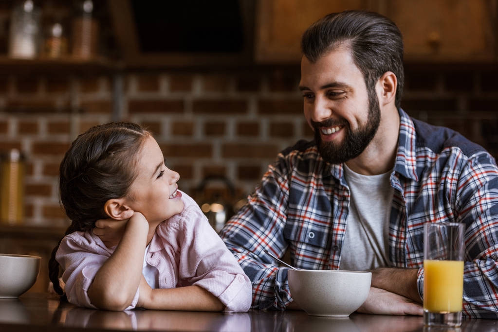 Šťastný otec s dcerou sedí u stolu s džusem a misky a při pohledu na sebe v kuchyni - Fotografie, Obrázek