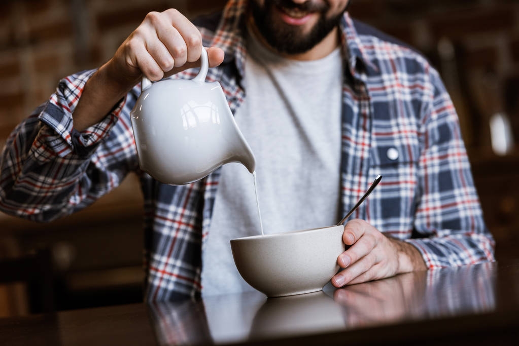 imagen recortada de hombre vertiendo leche en un tazón con bocadillos en la cocina
 - Foto, Imagen