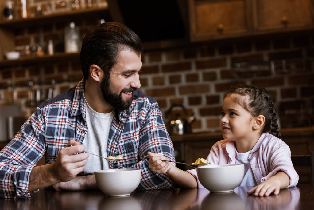 χαρούμενα πατέρας και κόρη κάθεται στο τραπέζι και τρώει σνακ με γάλα στην κουζίνα - Φωτογραφία, εικόνα
