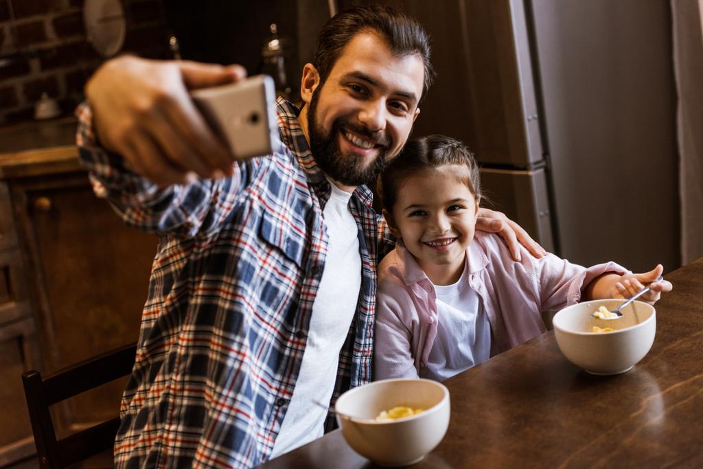 ευτυχής πατέρας με την κόρη που κάθεται στο τραπέζι με σνακ σε μπωλ και παίρνει selfie στην κουζίνα - Φωτογραφία, εικόνα