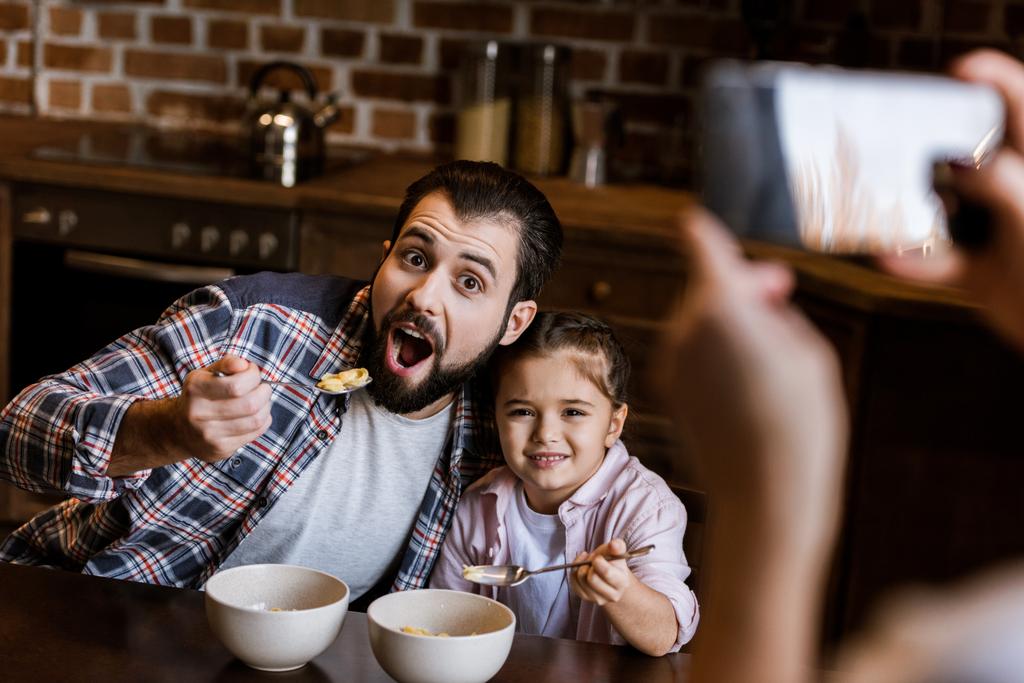 vrouw neemt de foto van vader met dochter zitten aan tafel en eten snacks uit kommen op keuken - Foto, afbeelding