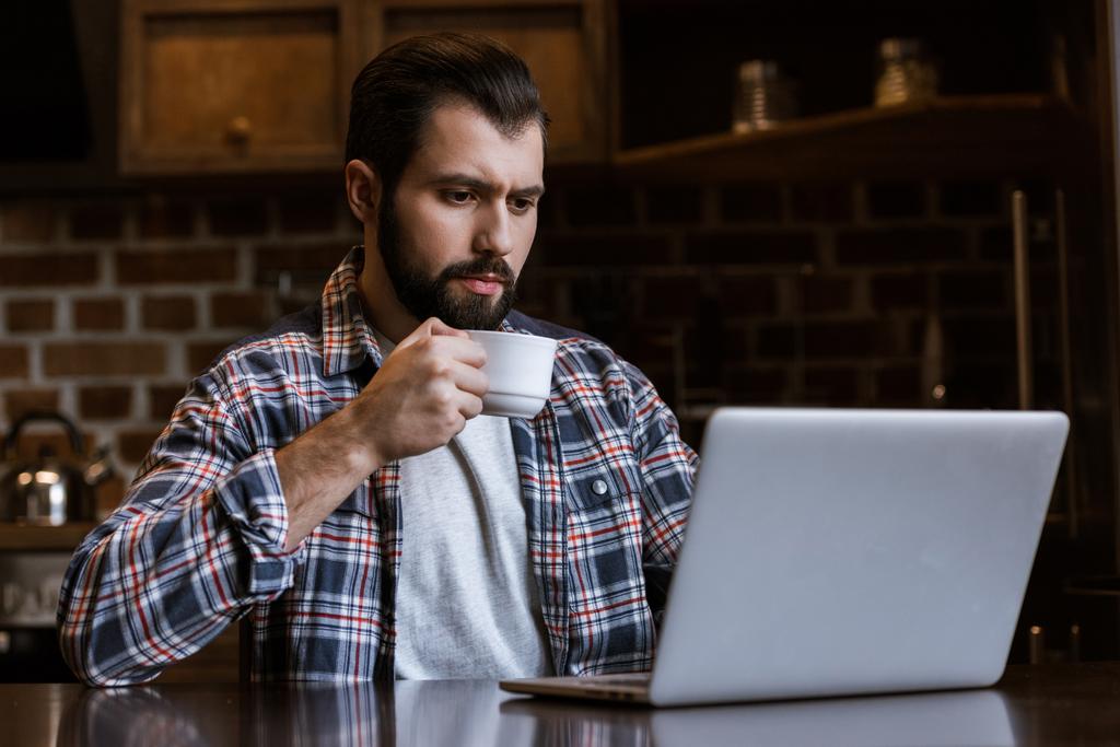 pohledný muž s šálek kávy sedí u stolu s notebookem - Fotografie, Obrázek