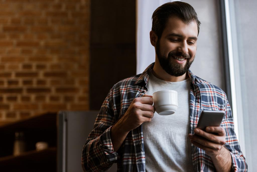 homme heureux tenant tasse de café et en utilisant smartphone à la maison
 - Photo, image