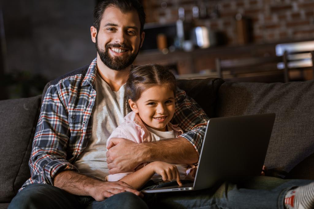 szczęśliwy ojciec z córką przytulanie na kanapie i komputera przenośnego - Zdjęcie, obraz