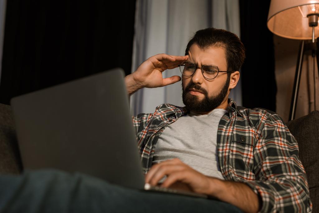 ένας άνθρωπος κουρασμένος, γυαλιά, κάθεται στον καναπέ με το laptop - Φωτογραφία, εικόνα