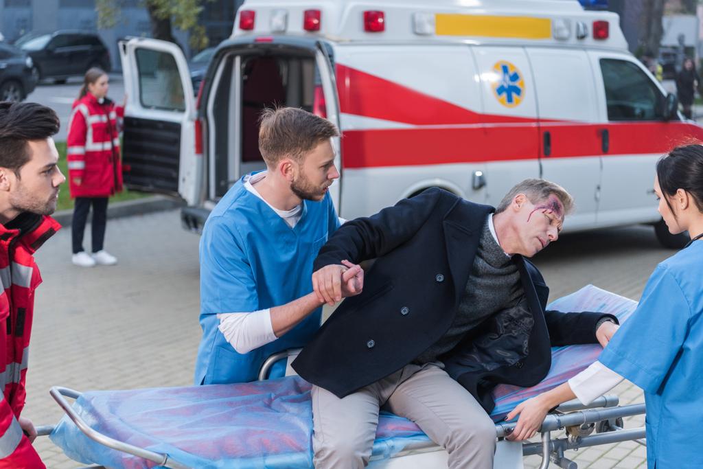 jonge paramedici helpen gewond man leugen neer op ambulance brancard - Foto, afbeelding